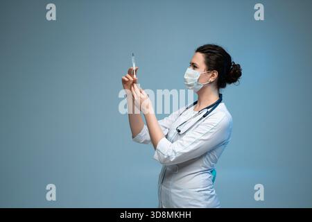 Die Krankenschwester untersucht die Spritze vor der Injektion mit Maske und Uniform in blauem klinischem Hintergrund Stockfoto