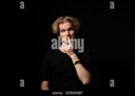 Denkender neugieriger Mann, der die Lippen berührt, unter Berücksichtigung von dunklem Hintergrund, ausdrucksstarkem Atelierporträt Stockfoto