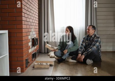Brennholz für den Heizofen von kaukasischen Männern und Frauen, die zusammen auf dem Boden in einem warmen rustikalen Innenraum mit natürlichem Licht sitzen. Winter Firewood Routine Vorbereitung und Beleuchtung eines Holzofens Stockfoto