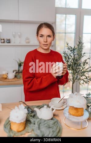 Frauenküche ostern, junge Frau in rotem Pullover, die Becher neben traditionellen Kulich-Kuchen auf einer Holztheke hält Stockfoto