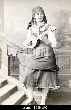 Florence St. John steht auf einer Studiotreppe und hält ein Banjo im Kostüm für die Titelrolle in Madame Favart im Strand Theatre, 1879. Ihr langer Rock, ihre Bluse und ihr Kopftuch spiegeln den theatralischen Stil dieser Zeit wider. Die gemalte Kulisse und die musikalische Requisite erinnern an viktorianisches Bühnenbild, Charakterdarstellung und das fotografische Flair der London Stereoscopic Company. Von einem späten viktorianischen bis frühen edwardianischen Original-Fotoalbum, das viele Albumenkarten und Visitenkarten enthielt und einen Einblick in die Stile und Moden der Zeit bot. Stockfoto