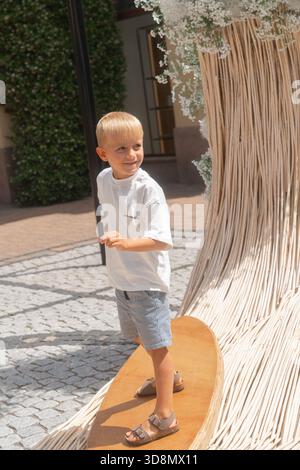 Ein Junge steht auf einer großen dekorativen wellenförmigen Holzskulptur in einem öffentlichen Außenbereich. Das helle Tageslicht unterstreicht die geschwungene Struktur Stockfoto