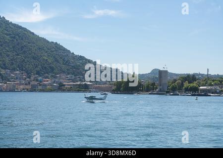 Ein kleines Boot fährt über einen ruhigen See, der von Bergen und einem Dorf am See umgeben ist. Eine ruhige Sommerszene mit sanftem Licht und freiem Raum Stockfoto