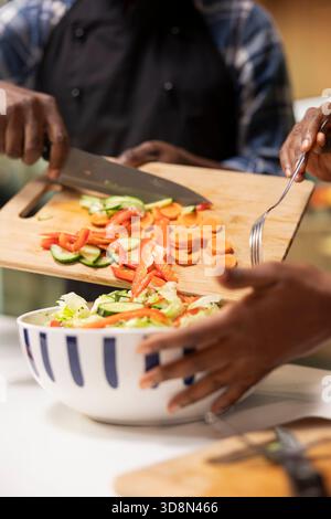 Nahaufnahme einer schwarzen Person, die frisch geschnittenes Gemüse in eine Schüssel gab, um leckeren Salat zu machen, und Karotten, Paprika und Gurken auf dem Schneidebrett hatte. Vegetarisches Paar bereitet zu Hause ein Mittagessen vor. Stockfoto