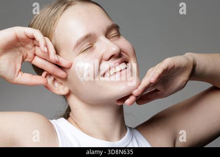Lächelnde Frau mit geschlossenen Augen und Herz der feuchtigkeitsspendenden Creme auf der Wange, die Freude und sanfte tägliche Gesichtspflege zeigt Stockfoto