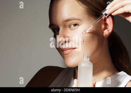 Nahaufnahme Porträt einer jungen Frau mit Glaspipette mit feuchtigkeitsspendendem Gesichtsserum, das natürliches Leuchten, saubere Haut und einfache Hautpflege zu Hause zeigt. Stockfoto