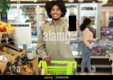Junger Mann zeigt Smartphone beim Einkaufen von Lebensmitteln im Supermarkt mit einer Frau, die in der Nähe stöbert Stockfoto