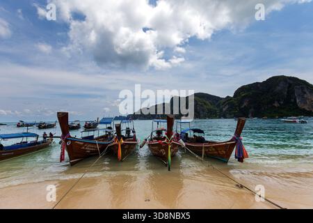 Blick auf traditionelle Longtail-Boote, die am Sandstrand ruhen, das türkisfarbene Wasser, das sanft gegen die Küste plätschert, Phi Phi Island, Krabi, Thailand. Stockfoto