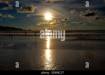 Blick auf die Sonne, die sich vom Wasser auf einem Strand spiegelt, mit Menschen, die den Sonnenuntergang in der Ferne genießen, Phuket, Phuket, Thailand. Stockfoto