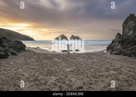 Der Blick auf eine Einzelfigur steht vor dem glitzernden Horizont, die Arme sind ausgestreckt, um das goldene Licht zu umarmen, eingebettet zwischen felsigen Felsvorsprüngen Stockfoto