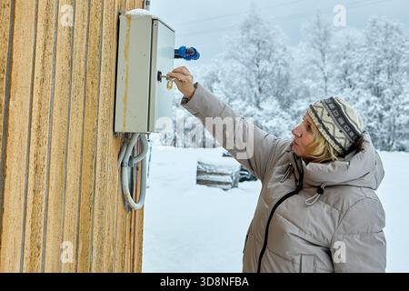 Stromzähler in Verbrauchereinheit, die an der Außenwand des Landhauses montiert ist, wird überprüft, ob die Verbraucherin die Tür mit dem Schlüssel im Winter öffnet. Stockfoto