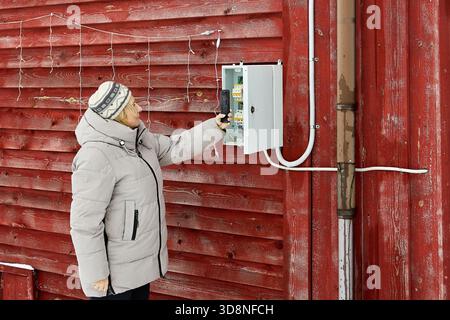 Das Sammeln von Zählerwerten der Energieverbraucher in warmer Kleidung richtet das Tablet an das elektrische Messgerät in der Verteilerwand an der Wand des Landhauses. Stockfoto