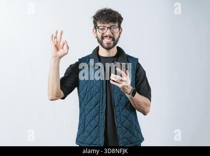 Lächelnder, gutaussehender Mann, der das Telefon hält und Gesten in Ordnung hält, isoliert. Attraktiver Mann in Brille mit Handygestik OK Stockfoto