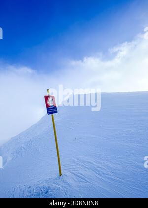 Ein Warnschild steht auf einem verschneiten Berghang unter einem hellblauen Himmel und schafft eine saubere Wintersicherheit und Outdoor-Abenteuer. Stockfoto