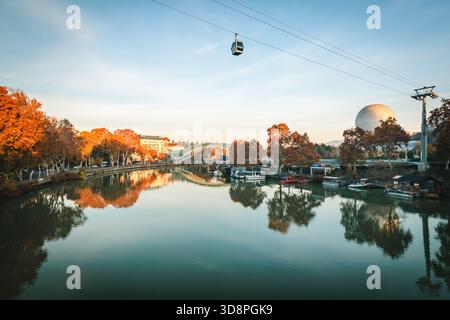 Goldener Herbstblick auf Tiflis mit der Friedensbrücke, die auf dem ruhigen Fluss Kura reflektiert, eingerahmt von farbenfrohen Blättern und Booten, die am Fluss du vertäut sind Stockfoto