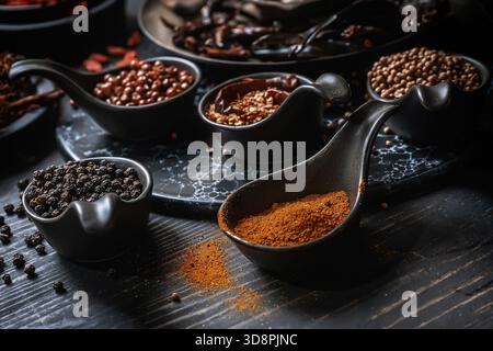 Verschiedene getrocknete Gewürze und Kräuter in Löffeln und Schüsseln auf dunklem Holzhintergrund. Stockfoto