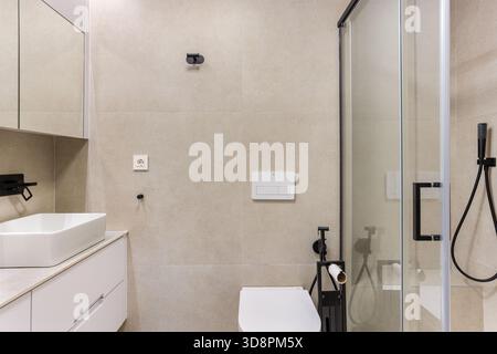 Modernes Bad mit Glasdusche, Waschbecken, beigefarbenen Fliesen und mattschwarzen Accessoires Stockfoto