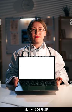 Die lächelnde Ärztin hält den Laptop mit leerem Bildschirm in der Hand, während sie in modernen Büros Nachtschicht arbeitet. Eine Therapeutin empfiehlt eine mobile App. Stockfoto