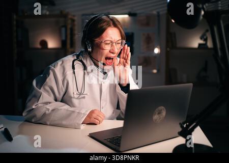 Weibliche Ärztin in Uniform tragen Headset schreiende Nachrichten und Geheimnis zur Kamera verwenden Laptop Computer sprechen auf Videoanruf mit Patienten im Krankenhaus während Stockfoto