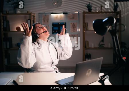 Wütend weibliche Ärztin in Uniform tragen Headset hat negative Emotionen Verwenden Sie Laptop-Computer, der in der Nacht mit dem Patienten im Krankenhaus spricht Stockfoto