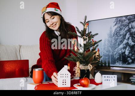 Die lächelnde Frau in einem Weihnachtsmann-Hut arrangiert gemütliche Weihnachtsdekorationen neben dem Weihnachtsbaum mit handgefertigten Ornamenten. Nostalgische Weihnachten, handgefertigte Ornamente Stockfoto