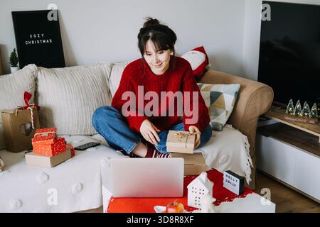Die junge Erwachsene Frau sitzt auf dem Sofa und hält eine kleine Geschenkbox, während sie ihren Laptop zum Einkaufen im Urlaub benutzt. Online-Shopping, saisonaler E-Commerce, Weihnachten Stockfoto