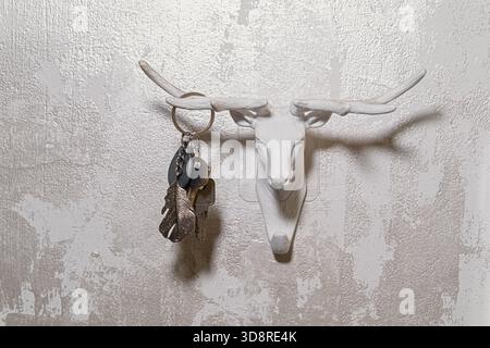 Dekorativer weißer Hirschkopf-Wandhaken mit Metallschlüsseln und Schlüsselanhänger, der an einer strukturierten Lichtwand hängt, symbolisiert die Organisation des Hauseingangs und ist modern eingerichtet Stockfoto