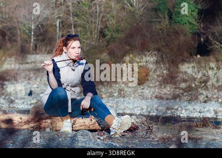 Rothaarige Frau in lässiger Kleidung sitzt im Kreuz auf Holzspieß, umgeben von Herbstlaub und weit entfernten Bäumen im Wald. Stockfoto