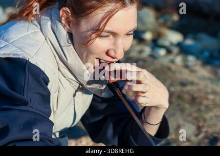 Aus nächster Nähe sehen Sie eine junge Frau, die einen Happen von gebratenem Marshmallow oder Wurst auf Spießen genießt und im Herbst zufrieden an den Felsen des Lagerfeuers lächelt. Stockfoto