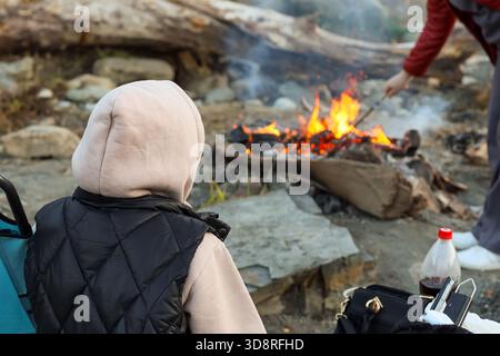 Eine Person in roter Jacke fügt einem Lagerfeuer Holz hinzu, während eine andere in Kapuze und Weste neben einem blauen Campingstuhl an einem felsigen Uferufer sitzt. Stockfoto