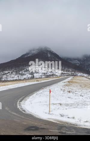 Gekrümmte Asphaltstraße, die zu einem Berg führt, der unter einem bewölkten Himmel mit Schnee und Nebel bedeckt ist Stockfoto