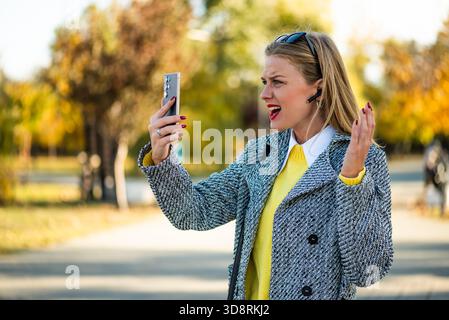 Wütende Geschäftsfrau, die draußen im Stadtpark steht und während eines stressigen Videoanrufs ins Smartphone ruft. Stockfoto