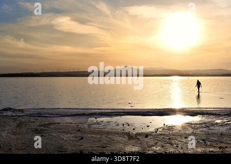 Silhouette eines Mannes, der während eines hellen Sonnenuntergangs am Larnaca Salt Lake, Zypern, mit Wolken am Himmel, ins Wasser geht. Stockfoto