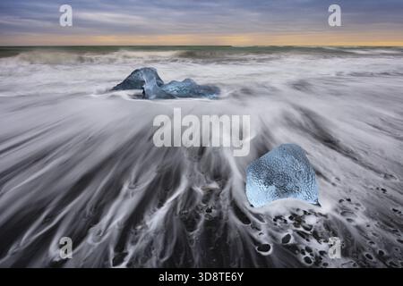Blick auf die eisigen Diamanten, die auf dem schwarzen Sandstrand verstreut sind, umspült von schaumigen Wellen unter einem pastellfarbenen Himmel, ein krasser Kontrast der kalten Schönheit, Diamond Beach, Stockfoto