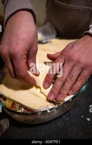 Nahaufnahme der Hände, die Tartlette mit Kartoffelschichten, Spachteln und Käse in einer Auflaufform in Cranves-Sales in Haute-Savoie in Auvergne-Rhone vorbereiten Stockfoto