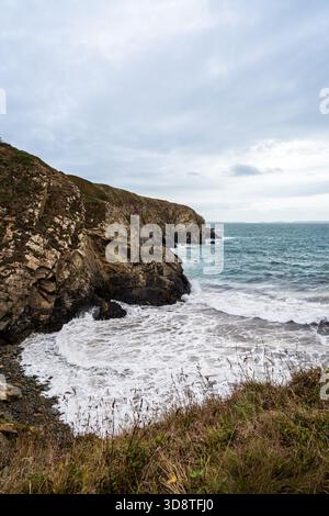 Blick auf zerklüftete Klippen treffen auf das turbulente Meer, weißen Schaum, der unter einem bewölkten Himmel gegen die dunklen Felsen kracht, St. Davids, Wales, Großbritannien. Stockfoto