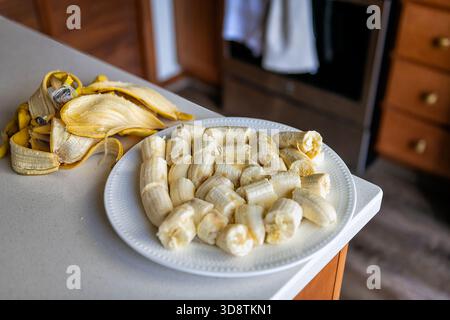 Bananen schneiden Stücke auf weißem Teller, Haufen von rohen, ungekochten geschälten Früchten mit Schalen zu Hause Küchentisch Theke Makronaht Stockfoto