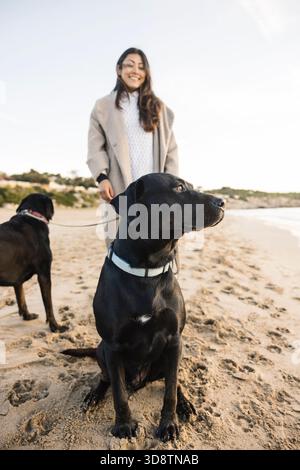 Frau lächelt, während sie zwei Hunde an einem sonnigen Strand spaziert Stockfoto