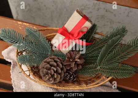 In einem rustikalen Korb befindet sich eine kleine Geschenkbox mit einem leuchtend roten Band, umgeben von immergrünen Zweigen und Tannenzapfen, die ein gemütliches, festliches Herzstück für die Weihnachtsdekoration und Geschenke bilden. Hochwertige Fotos Stockfoto