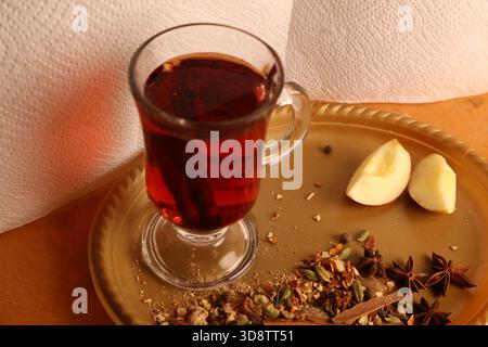 Eine gemütliche Szene mit einem Glas rotem Tee auf einem beigefarbenen Teller, Apfelscheiben und verstreuten Gewürzen wie Sternanis und Nelken. Perfekt für Herbst, Komfort und Wärme im Urlaub. Hochwertige Fotos Stockfoto