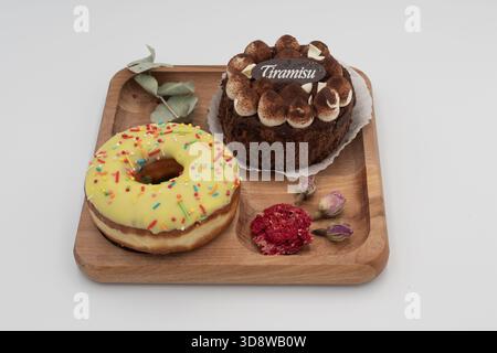 Modernes Dessert-Set mit gelbem Donut, Tiramisu-Kuchen und getrockneten Blumen auf Holztablett auf weißem Hintergrund Stockfoto
