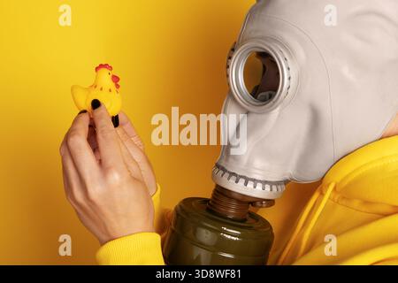Nahaufnahme einer Person in einer Gasmaske, die ein gelbes Gummihuhn vor einem leuchtend gelben Hintergrund untersucht. Konzeptionelles und kreatives Image. Stockfoto
