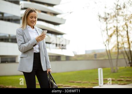 Eine Frau in einem grauen Blazer steht draußen und lächelt, während sie auf ihr Smartphone schaut. Sie befindet sich in der Nähe zeitgenössischer Gebäude mit grünem Gras und Bäumen Stockfoto