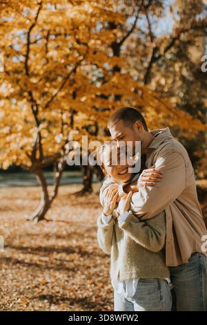 Eine Frau lächelt weit, während sie von einem Mann umarmt wird, inmitten lebendiger Herbstblätter in einem sonnendurchfluteten Park. Sie genießen einen freudigen Moment zusammen. Stockfoto