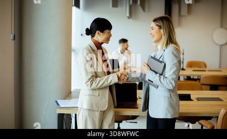 In einem modernen Büro mit stilvollem Dekor schütteln zwei professionelle Frauen nach einem erfolgreichen Meeting warm die Hände. Ein Mann sitzt im Hintergrund, fokussiert Stockfoto