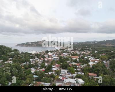 Malerischer Blick auf ein Küstendorf, umgeben von üppigem Grün und sanften Hügeln, mit einem ruhigen Ozean im Hintergrund unter einem bewölkten Himmel. Stockfoto