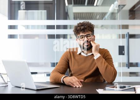 Bärtiger männlicher Mitarbeiter, der starke Zahnschmerzen spürt, seine Wange mit der Hand berührt, während er an einem Laptop in einem modernen Co-Working-Raum arbeitet, leidet unter Beschwerden und Erschöpfung Stockfoto