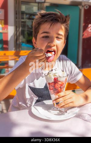Nahaufnahme eines fröhlichen Kindes, das an einem hellen Sommertag im Freien einen köstlichen Erdbeer-Eisbecher mit Schlagsahne genießt. Frohe Ferien und Reisen im Geheimen Stockfoto