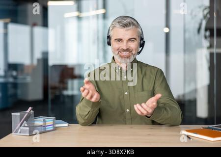 Ein reifer, bärtiger Geschäftsmann lächelt und spricht während eines virtuellen Meetings oder Webinars von einem modernen Büro aus in ein Headset, der sich aus der Ferne mit Kundensupport oder Coaching befasst Stockfoto