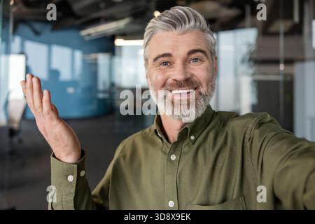 Lächelnder, reifer Geschäftsmann, der eine Videokonferenz anruft oder ein Video aufnimmt, mit freundlicher Geste winkt und in einer modernen Büroumgebung direkt auf die Kamera blickt Stockfoto